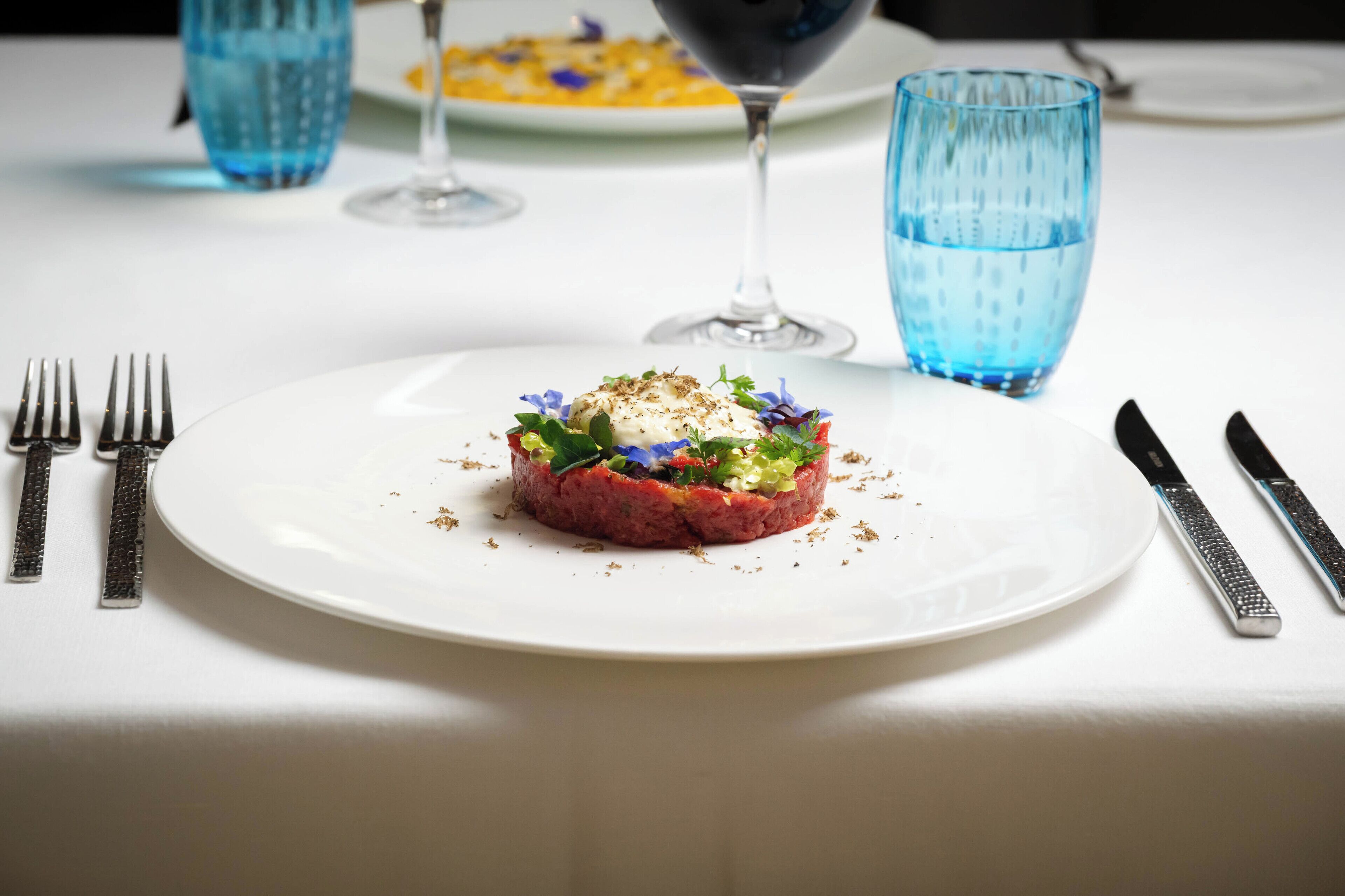 Une assiette blanche contenant un plat à base de viande, accompagnée d’une fourchette, d’un couteau et de deux verres bleus, le tout posé sur une nappe blanche.
