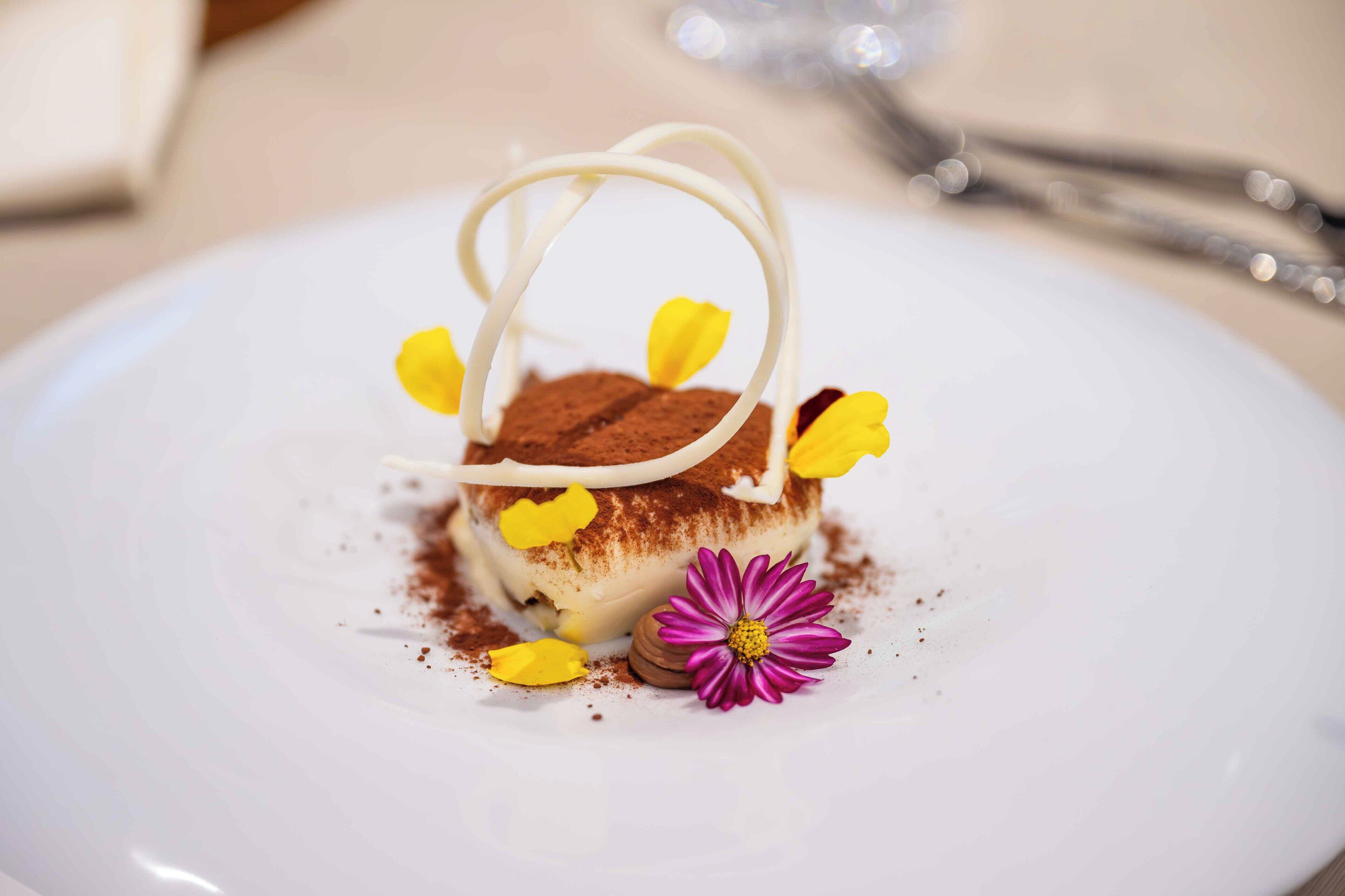 Un dessert su un piatto bianco, con un fiore viola e petali gialli.