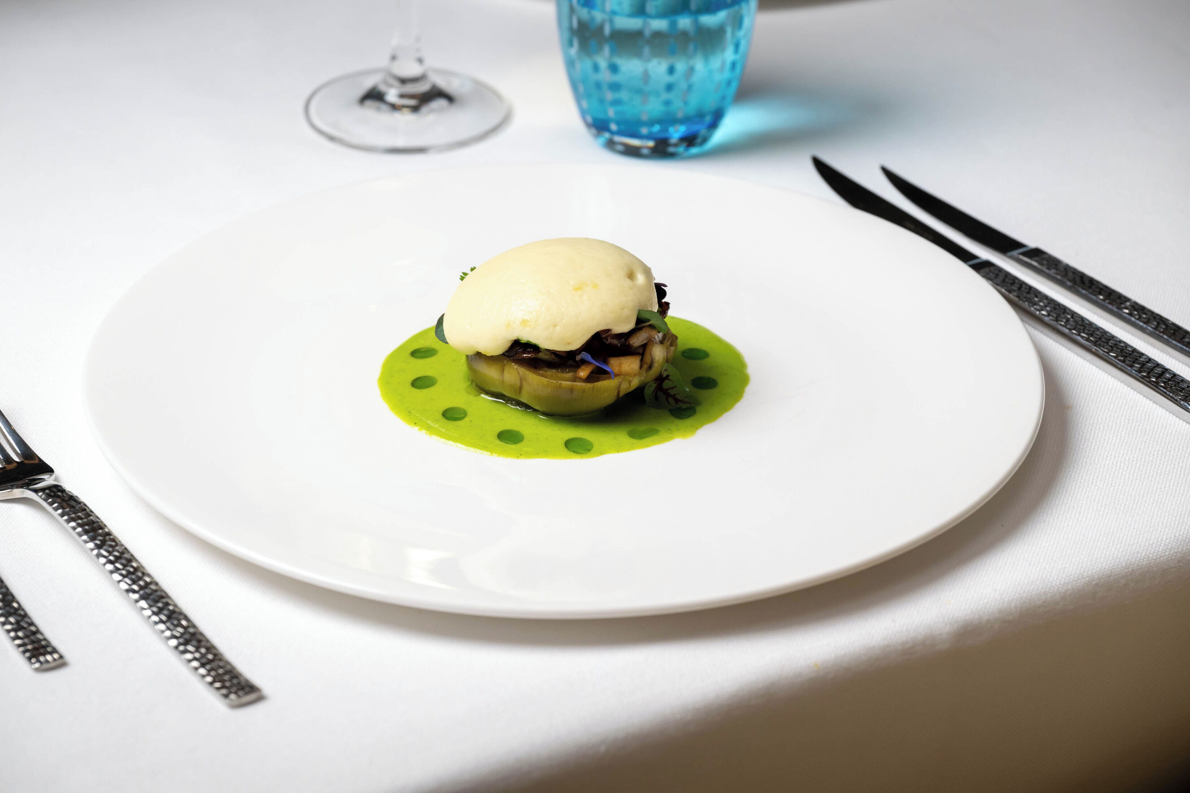 Une assiette blanche sur laquelle est disposé un dessert, elle-même posée sur une assiette verte à pois, accompagnée de couverts et d’un verre d’eau.