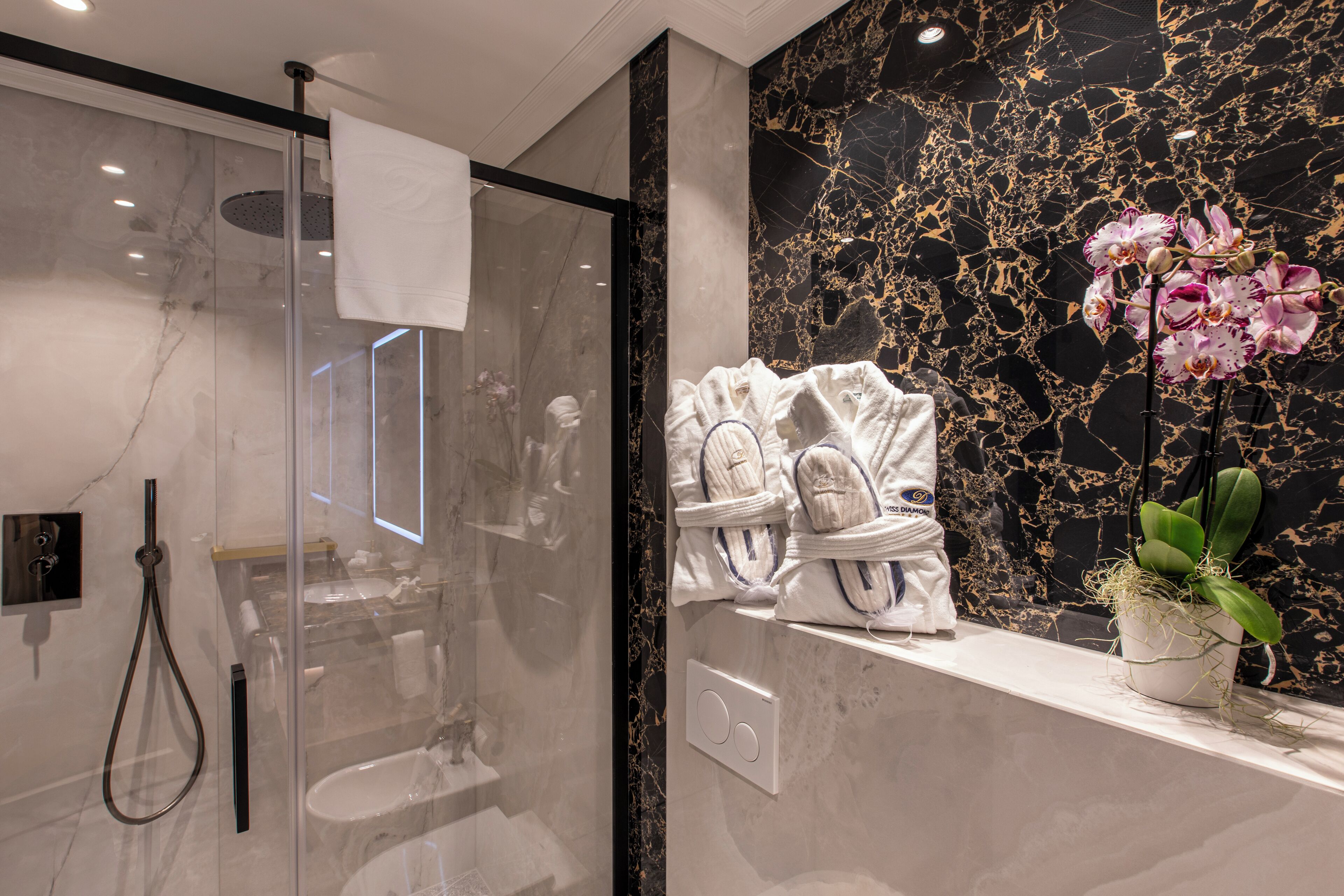 A modern bathroom with a glass-enclosed shower, marble walls, and a shelf with towels and a potted orchid.