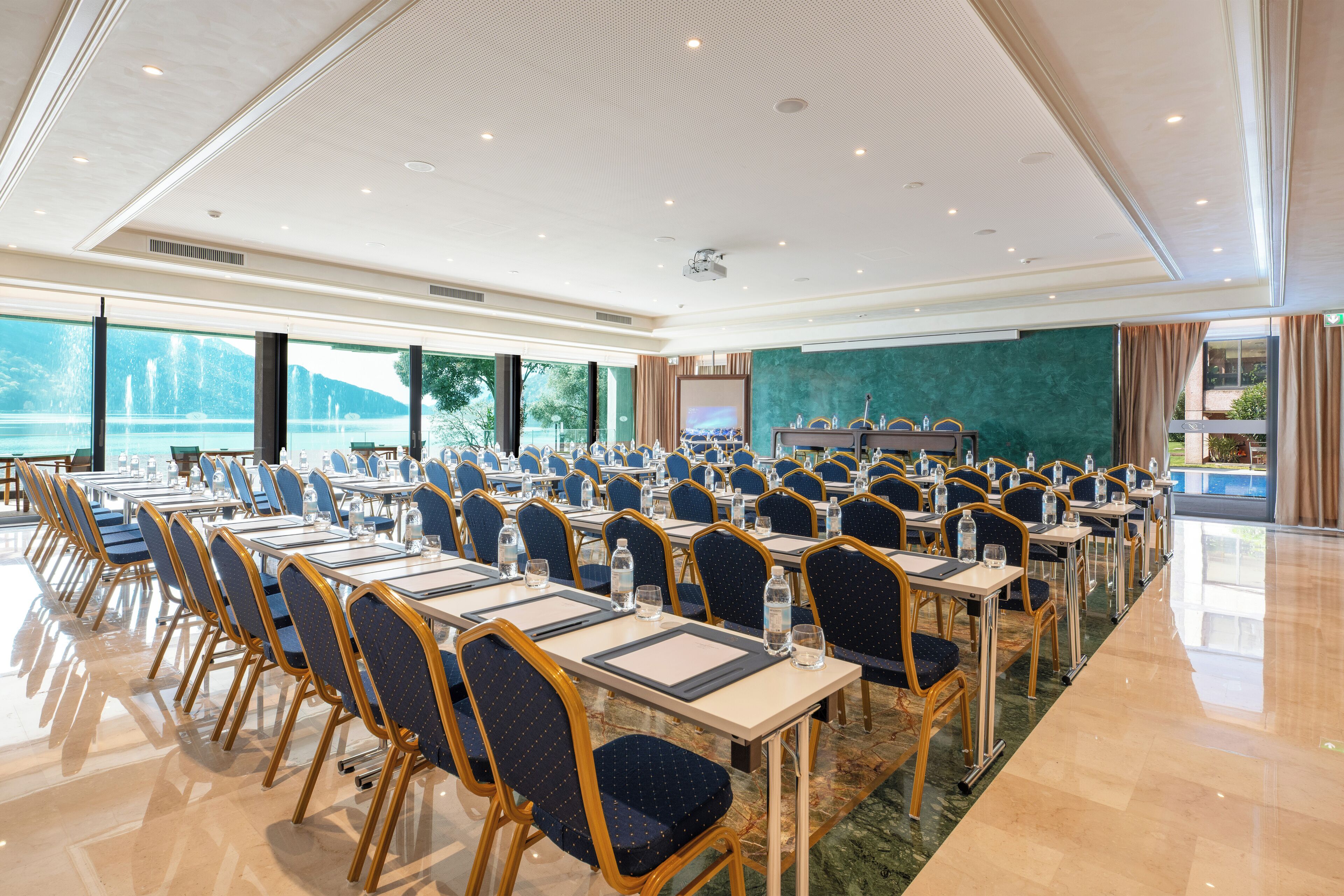 Un salón de conferencias con filas de mesas y sillas, grandes ventanales con vista a árboles y agua, y un escenario con un atril.