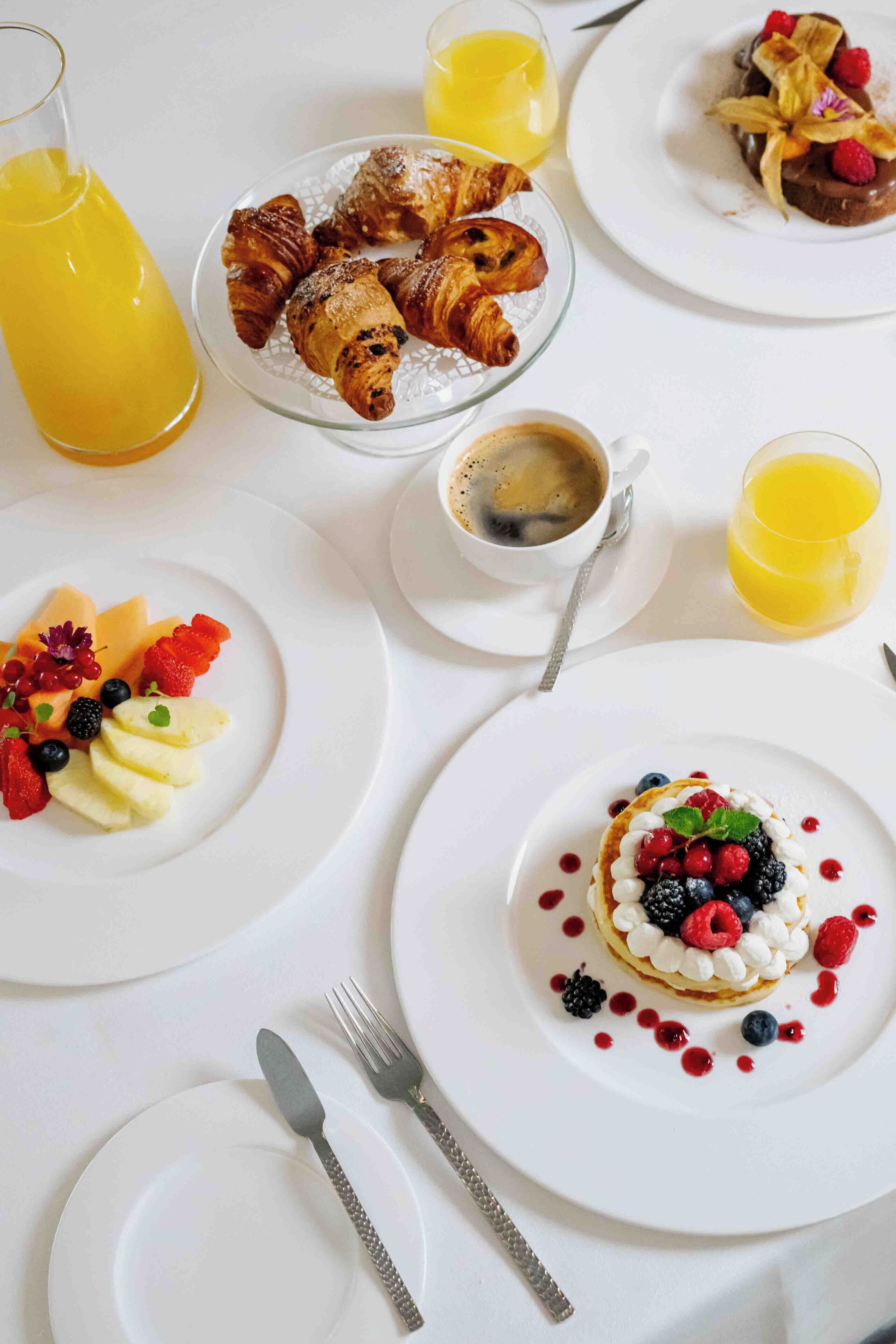 A breakfast setting with croissants, fruit, and a dessert on plates, along with a glass of orange juice and a cup of coffee.