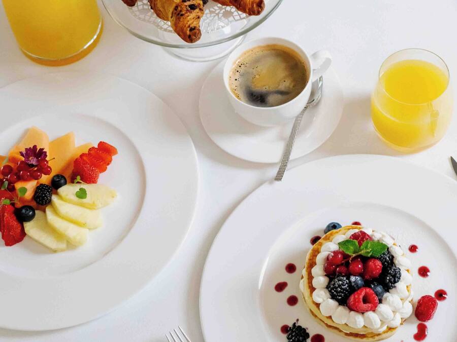 Un desayuno con cruasanes, fruta y un postre en platos, junto con un vaso de jugo de naranja y una taza de café.