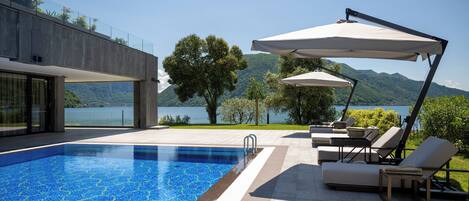 A modern outdoor pool area with lounge chairs and umbrellas, overlooking a lake and mountains.