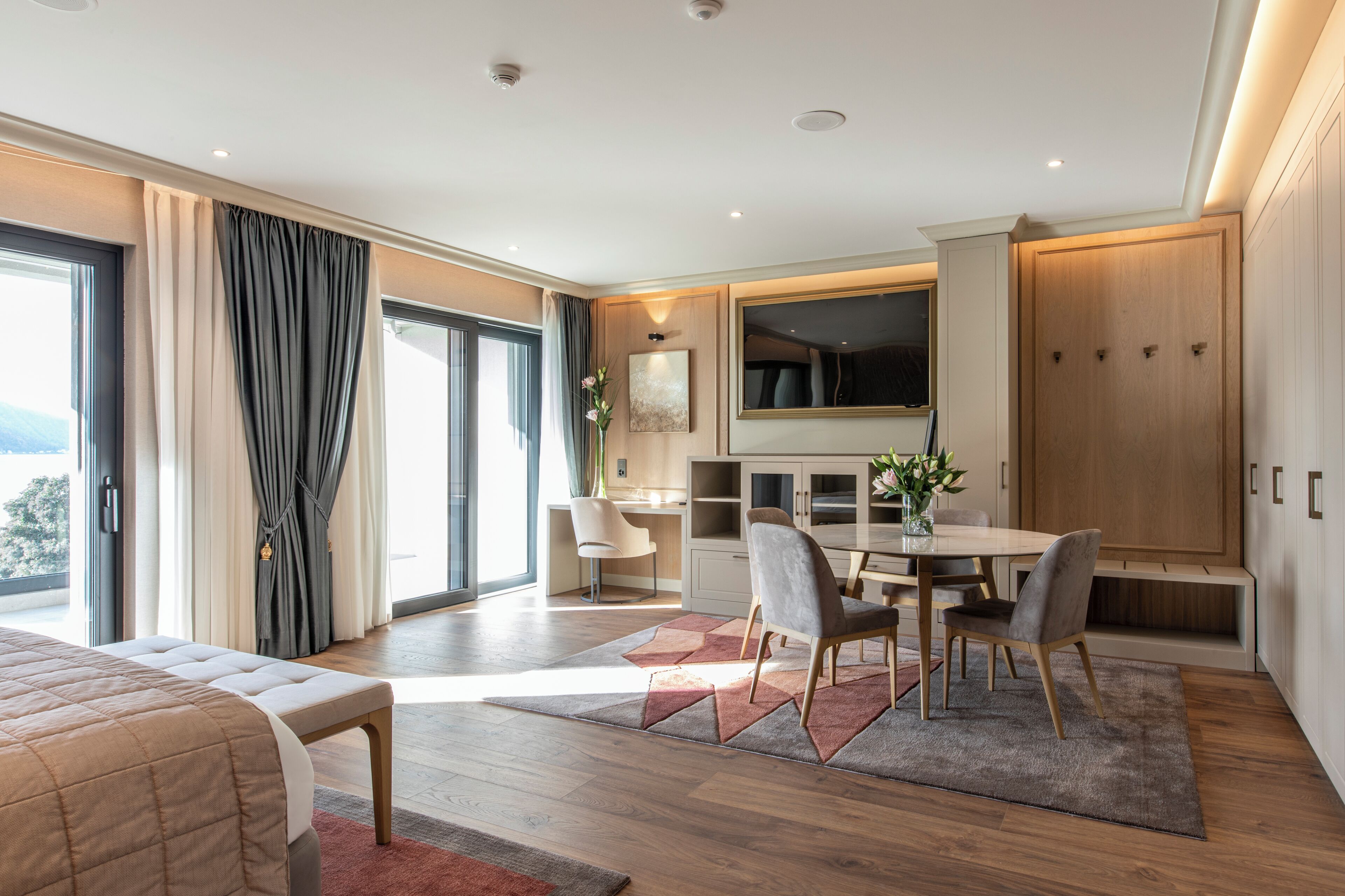 Ein modernes Hotelzimmer mit einem großen Bett, einem Essbereich mit Tisch und Stühlen, einem an der Wand montierten Fernseher und einem großen Kleiderschrank.