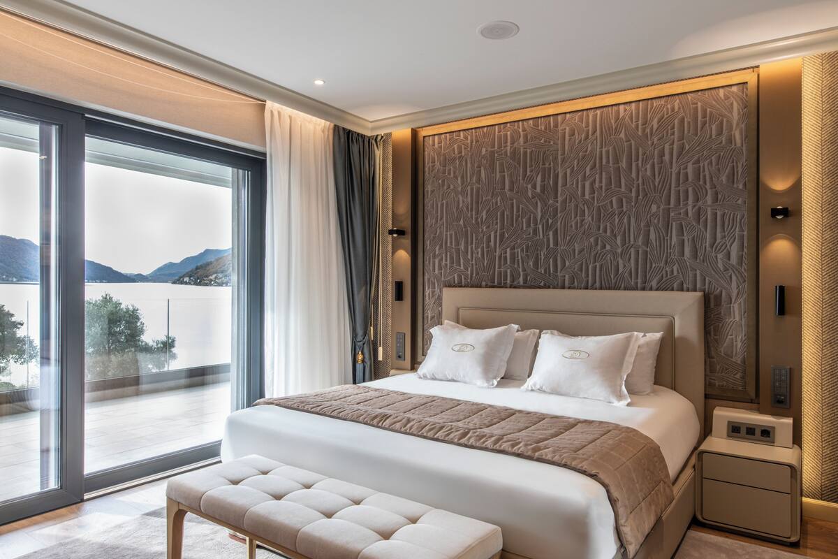 A modern hotel room with a large bed, a bench, a view of the outdoors, and a decorative wall panel.