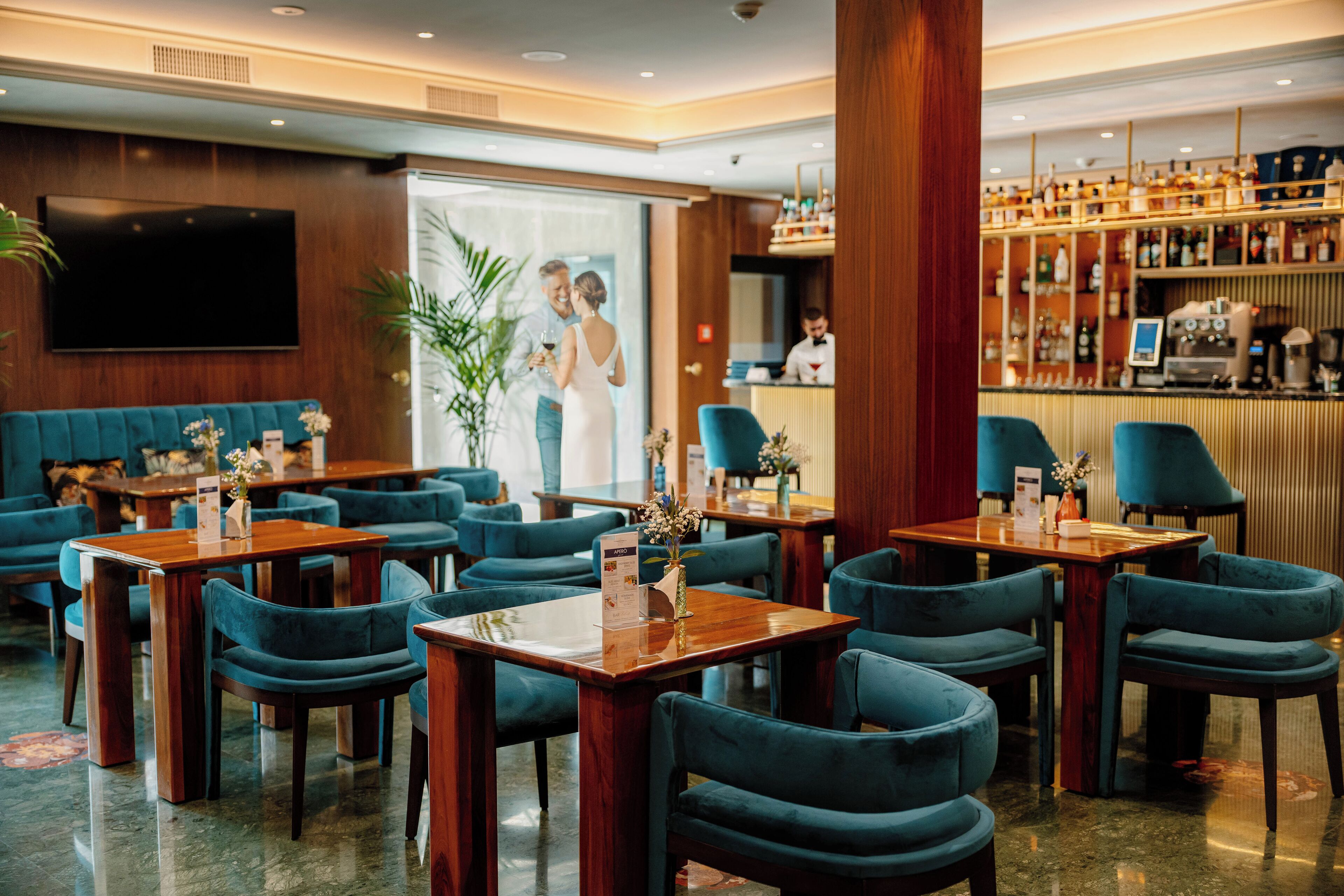 Een hotellobby met houten tafels en blauwe stoelen, een bar met flessenkasten, en een grote flatscreen televisie aan de muur.