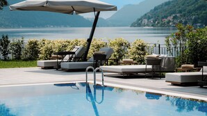 A poolside area with lounge chairs and umbrellas, a clear view of the lake, and mountains in the background. - Swiss Diamond Boutique Hotel La Romantica (Melide)