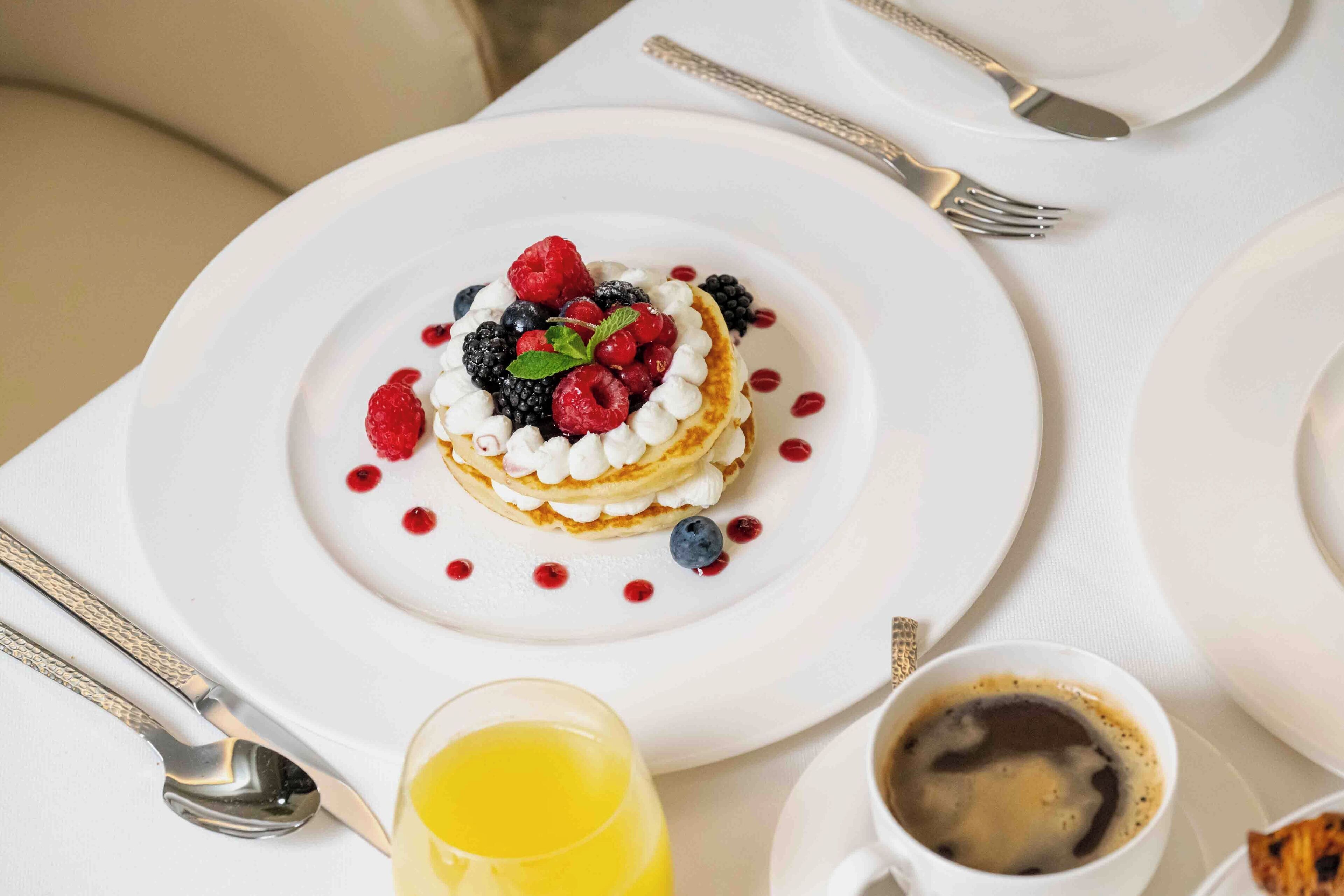 Mesa de café da manhã com panquecas cobertas de frutas vermelhas e chantilly, um copo de suco de laranja e uma xícara de café sobre uma toalha de mesa branca.