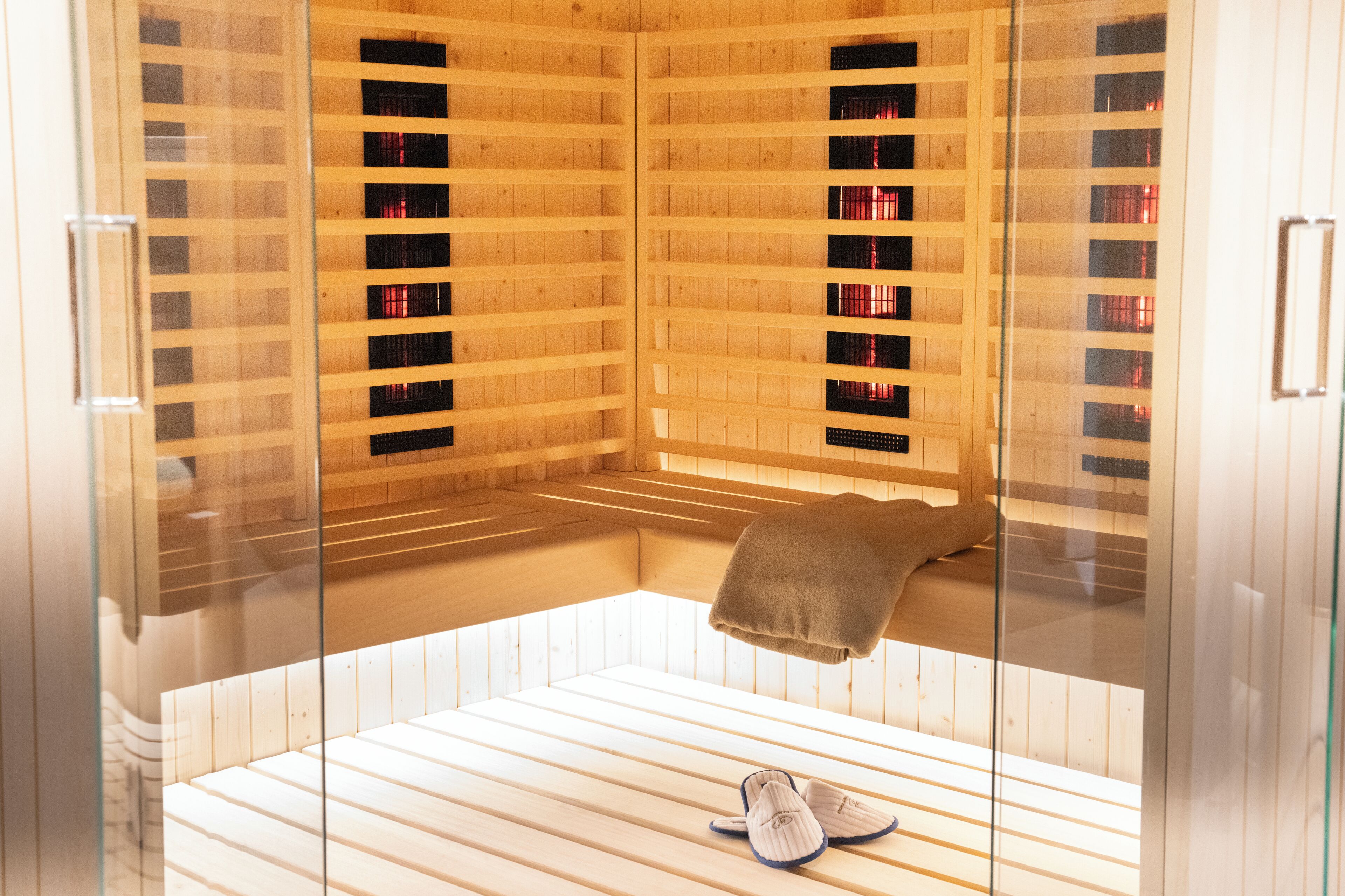 A sauna with glass walls, wooden benches, and a bench with slippers.