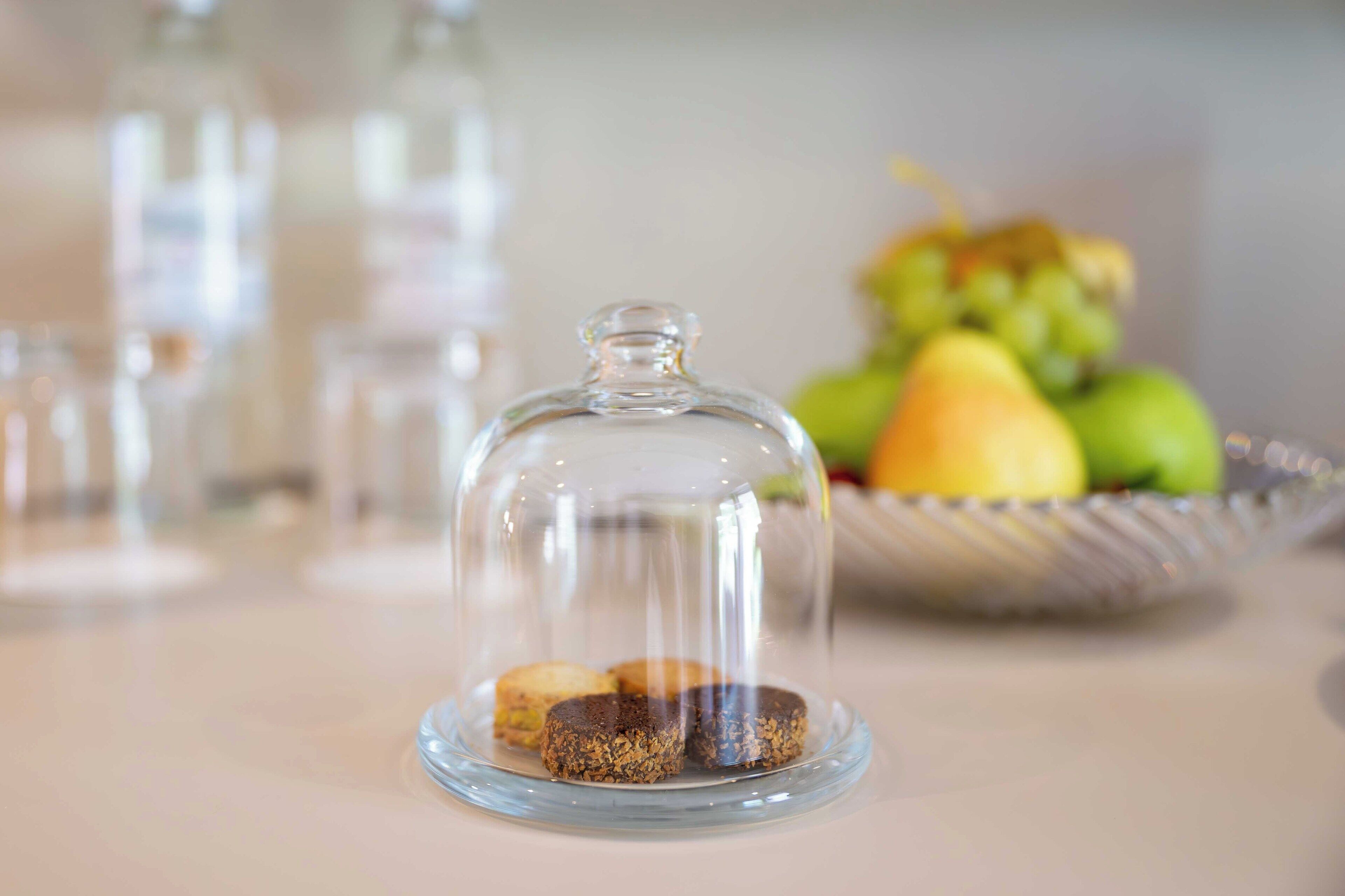Una cupola di vetro sopra un piatto con tre biscotti, e in secondo piano una ciotola di frutta.