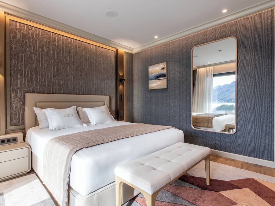 Een moderne slaapkamer met een groot bed, nachtkastjes, een bankje, een spiegel en uitzicht op de bergen door het raam.