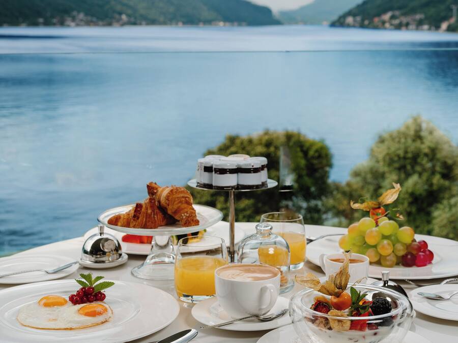 A breakfast table with croissants, fruit, and coffee, overlooking a lake with mountains in the background.