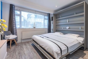 Une chambre à coucher moderne avec un lit, une fenêtre avec des rideaux, un radiateur, une chaise et une étagère ornée d’objets décoratifs.