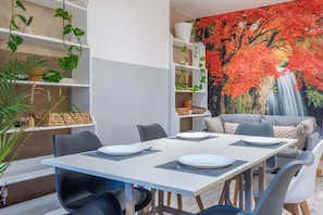 Un espace repas avec une table dressée pour quatre personnes, entourée de chaises, avec un canapé et une fresque murale en arrière-plan.