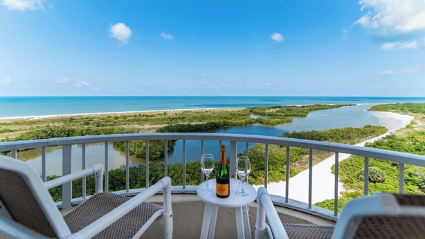 Ein Balkon mit Blick auf eine Küstenlandschaft, ein Tisch mit Flasche und Gläsern sowie zwei Stühle.