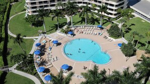 Ein Hotel mit einem großen Pool, umgeben von Liegestühlen und Sonnenschirmen, Palmen und einem mehrstöckigen Gebäude im Hintergrund.