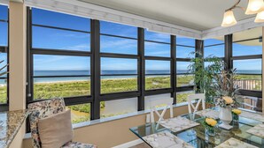 Ein Zimmer mit großen Fenstern und Blick auf eine Küstenlandschaft, ein mit weißem Geschirr gedeckter Esstisch und ein Blumentopf am Fensterbrett.