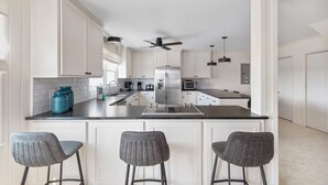 Uma cozinha moderna com armários brancos, uma ilha central com banquetas, um ventilador de teto e iluminação pendente.