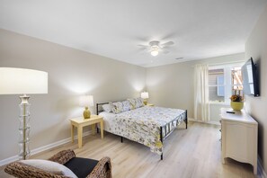 A bedroom with a bed, bedside tables, a lamp, a ceiling fan, a window with curtains, and a TV mounted on the wall. - Haney's Penthouse - Tri-plex, 4-bedroom (Pawleys Island)