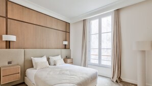 A modern hotel room with a large bed, bedside tables, a nightstand, and a window with curtains.