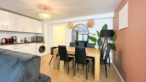 Cocina compacta con lavadora, microondas y cafetera. Hay un comedor con mesa y sillas, y una planta grande en la esquina.