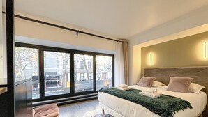 Habitación de hotel moderna con un ventanal grande, una cama con almohadas, una mesita de noche y vistas a la calle.