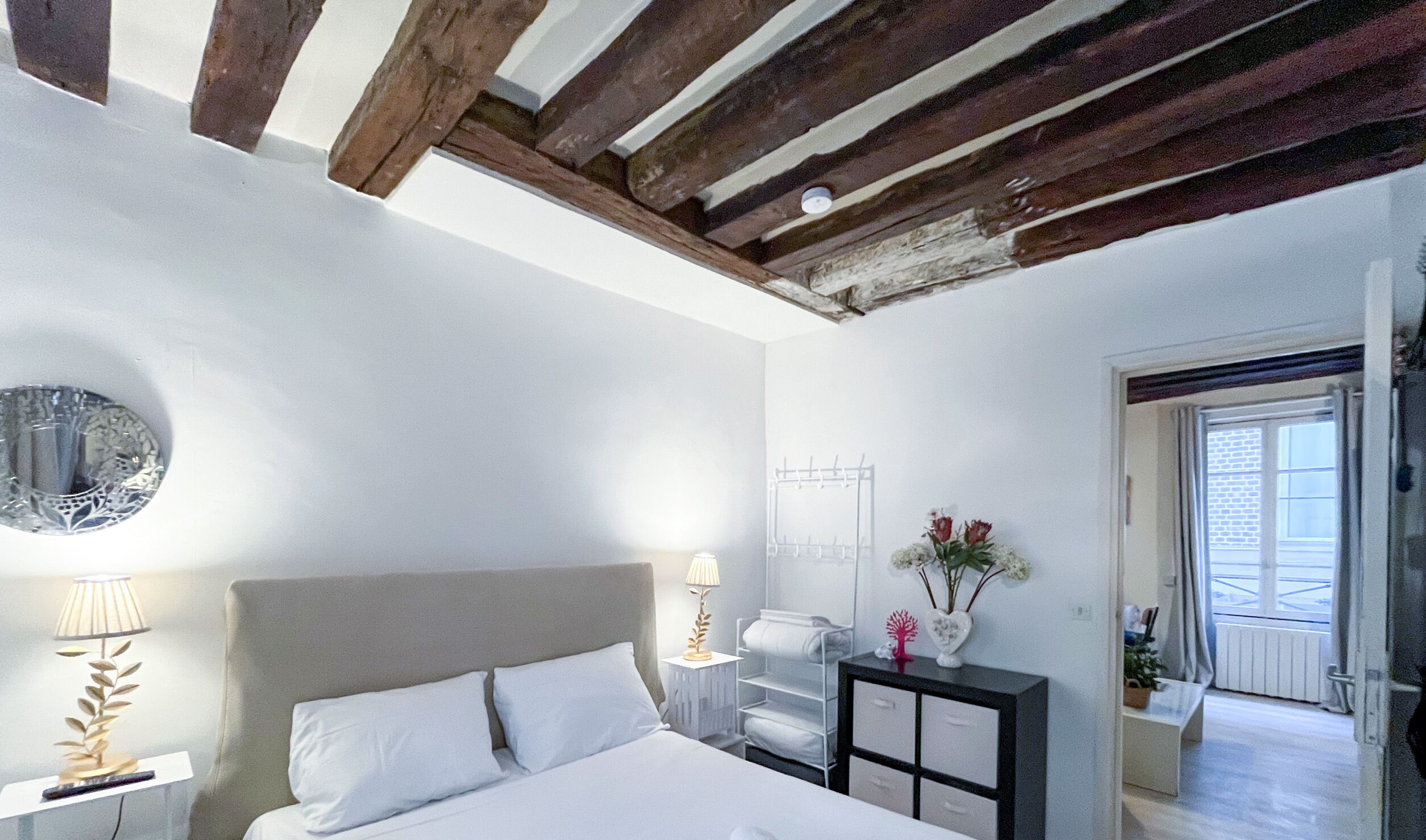 Quarto com cama, mesas de cabeceira, abajur, relógio na parede, radiador e janela.
