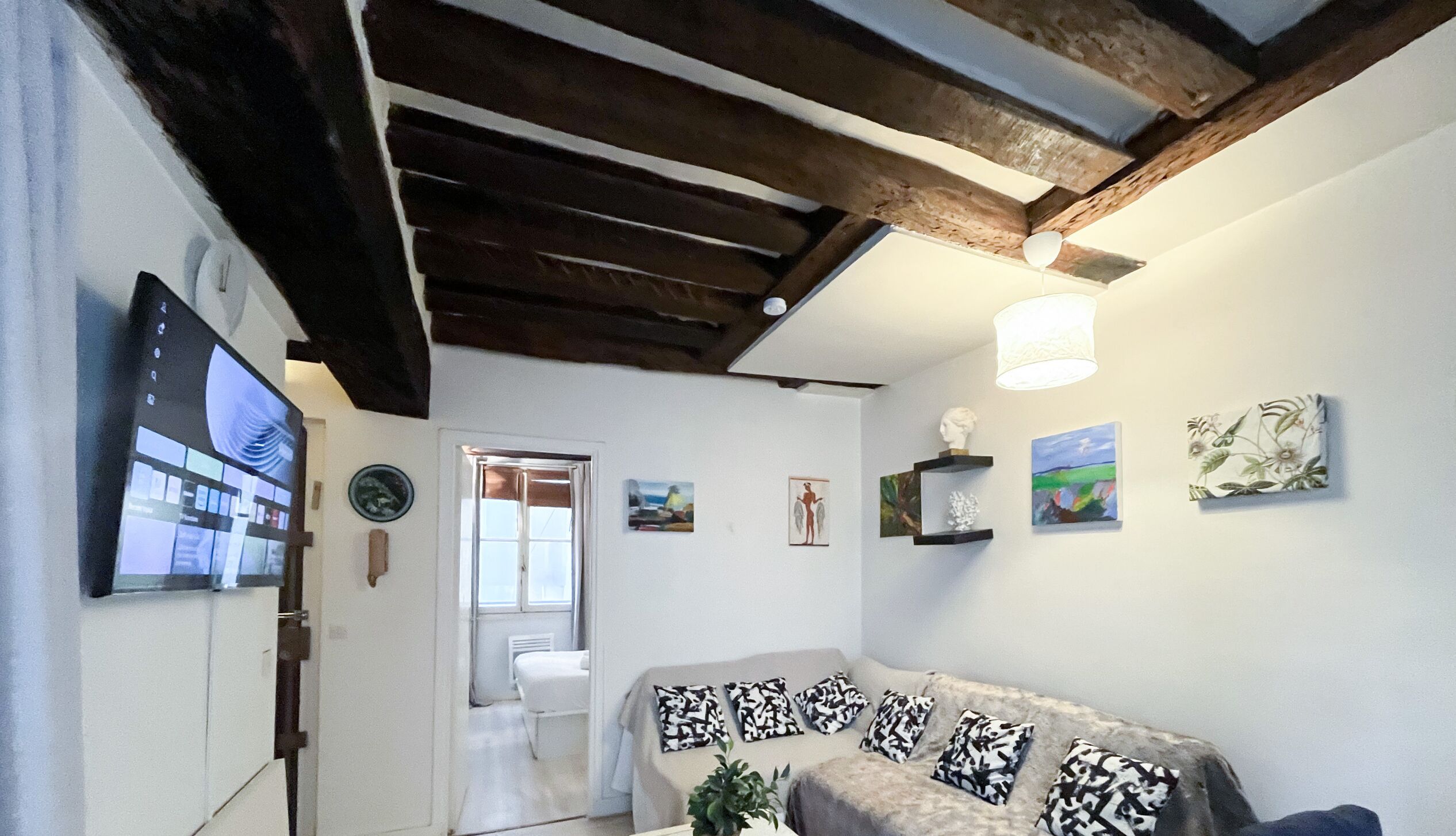 Een woonkamer met een bank, kunstwerken aan de wanden, een borstbeeld op een plank en een plafond met zichtbare balken.