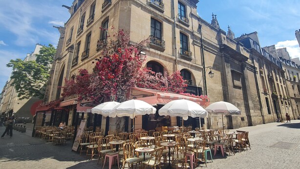 Un café avec des tables en terrasse, un bâtiment en pierre doté de fenêtres en arc de cercle et un arbre aux fleurs roses.