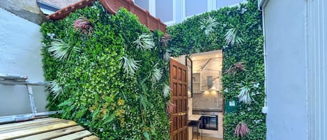 A narrow indoor space with a wooden bench, a door leading to a kitchen area, and walls covered with green foliage.