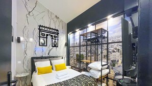 Une chambre moderne avec vue sur la ville, un lit avec une literie blanche et jaune, et une œuvre d’art murale originale.