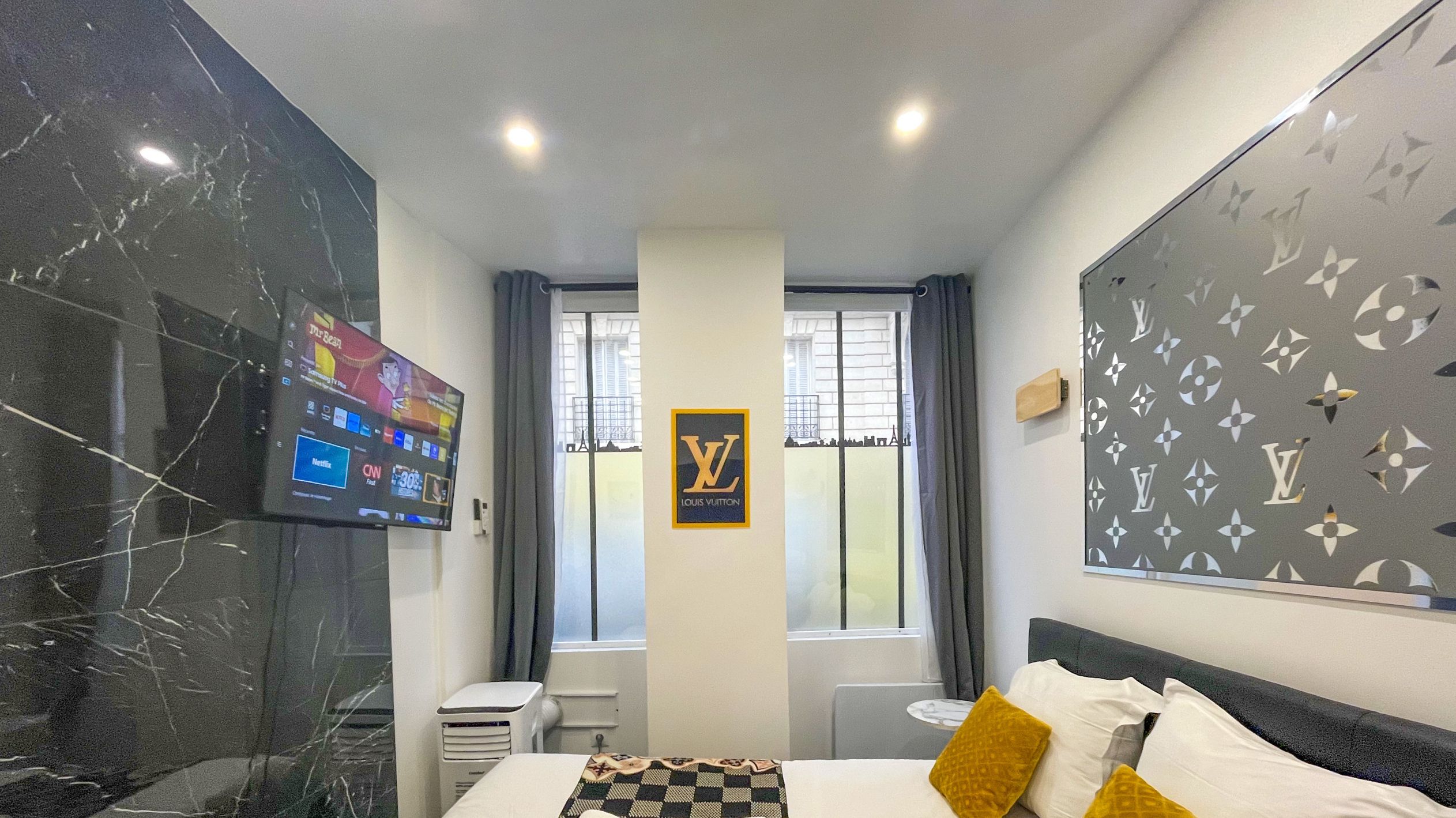 A hotel room with a bed, a TV mounted on the wall, a window with curtains, and a Louis Vuitton sign on the wall.