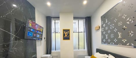 Una habitación de hotel con una cama, un televisor montado en la pared, un ventanal con cortinas y un letrero de Louis Vuitton en la pared.