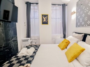 A hotel room with a bed, two pillows, a towel, a wall-mounted sign, a window with curtains, and a wall with a pattern.