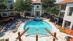 A swimming pool with lounge chairs and a central fountain, surrounded by buildings with balconies and trees. - Boulevard Stays 1BR King Suite Pool, Gym & Parking 03 (Irving)