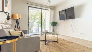 A modern living room with a sofa, a glass coffee table, a floor lamp, a TV mounted on the wall, and a large window with a view of greenery outside. - Boulevard Stays 1BR King Suite Pool, Gym & Parking 03 (Irving)