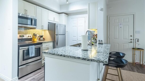 A modern kitchen with white cabinets, stainless steel appliances, a granite countertop, and a bar stool. - Boulevard Stays 1BR King Suite Pool, Gym & Parking 03 (Irving)
