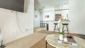 A modern living room with a flat-screen TV mounted on the wall, a round marble coffee table with decorative items, a kitchen area with a microwave and bar stools, and a large mirror reflecting the space. - Boulevard Stays 1BR King Suite Pool, Gym & Parking 03 (Irving)