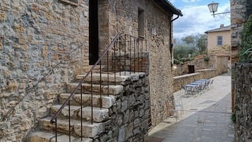 A stone building with a staircase leading to a door, a paved courtyard with outdoor seating, and a lamp post.