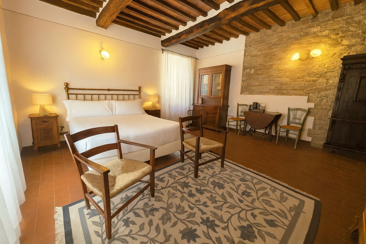 Un dormitorio con cama, dos sillas, una mesita de noche, un armario de madera y una pared de piedra.