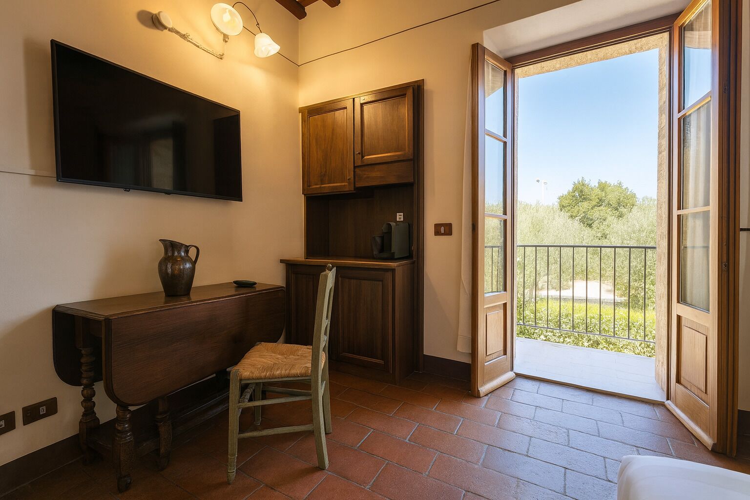 A room with a wooden desk, chair, and cabinets, a television, and a balcony with a view of greenery.