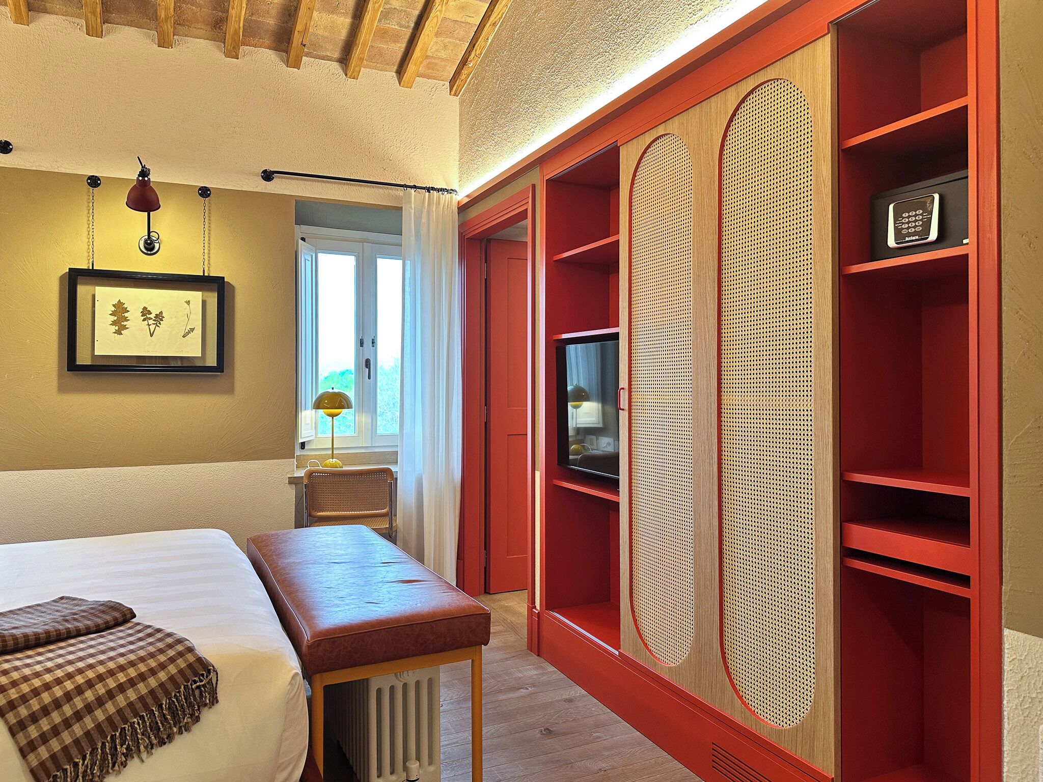 Ein Hotelzimmer mit einem Bett, einer Sitzbank, einem Fernseher und einem roten Kleiderschrank mit Luftlöchern.