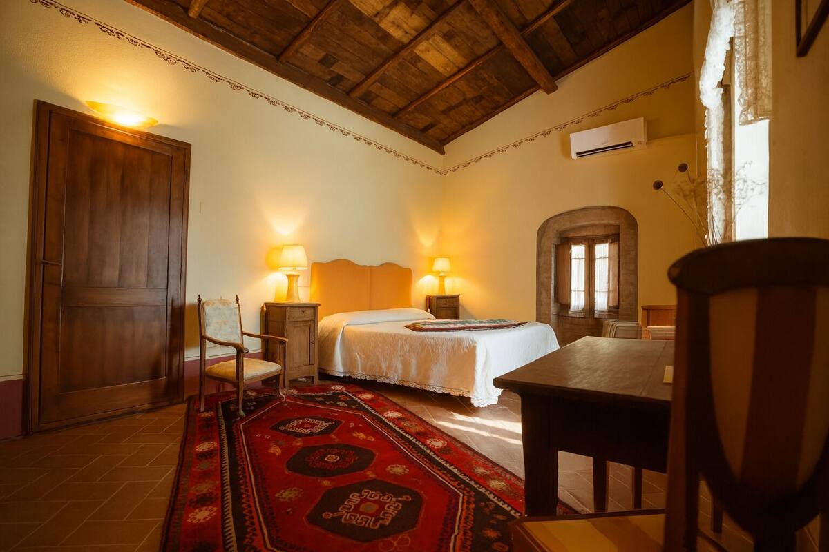 A bedroom with a wooden ceiling, a bed with white linens, two bedside lamps, a red patterned rug, and a wooden door.