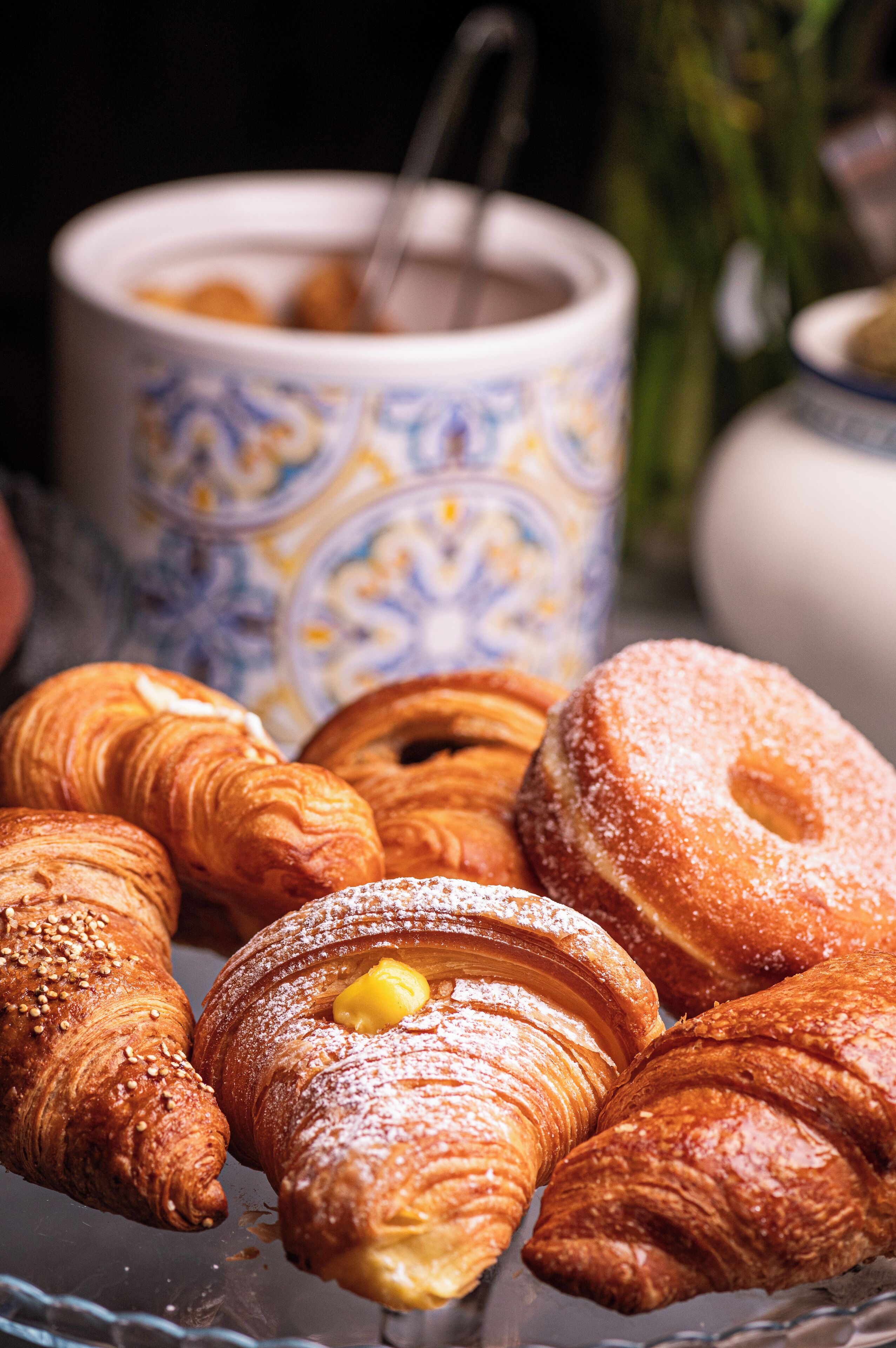 Ett urval av bakverk, inklusive croissanter och ett bakverk täckt med socker, arrangerade på en glasskiva med en dekorativ mugg i bakgrunden.