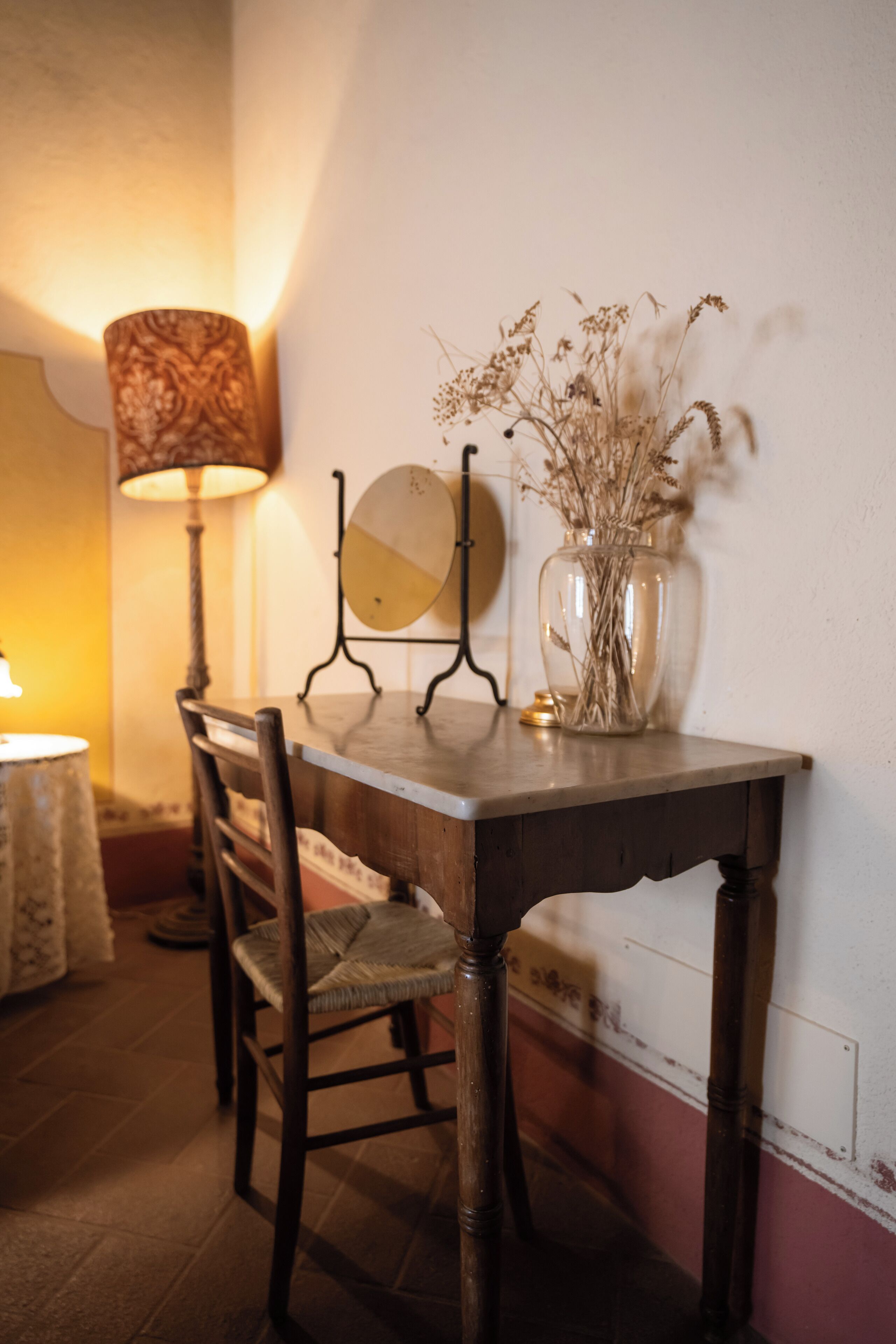 A dining table with chairs, a lamp, a vase with dried flowers, and a wall-mounted mirror.