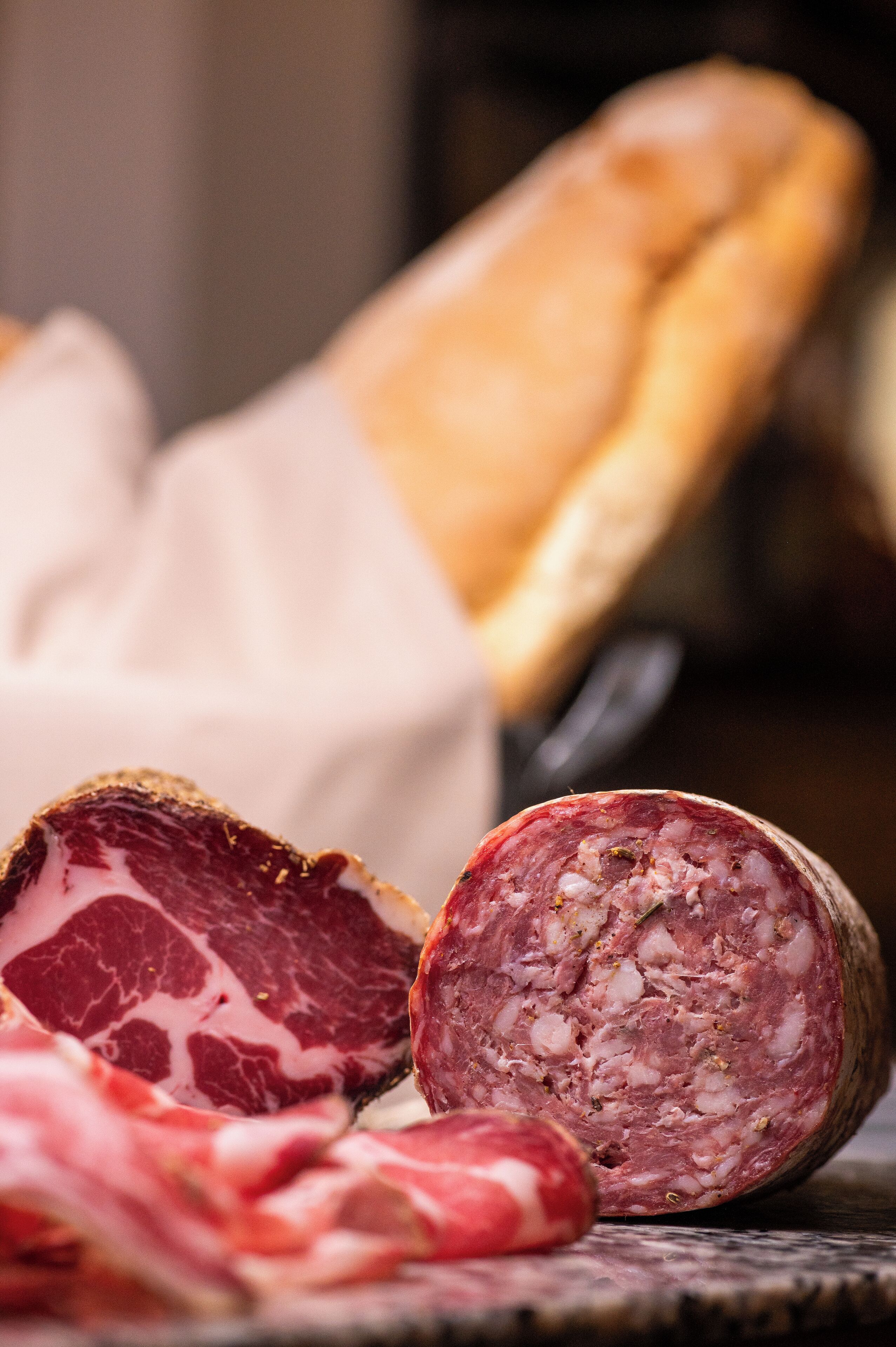 A close-up of sliced cured meat with a crusty exterior, a knife, and a loaf of bread in the background.