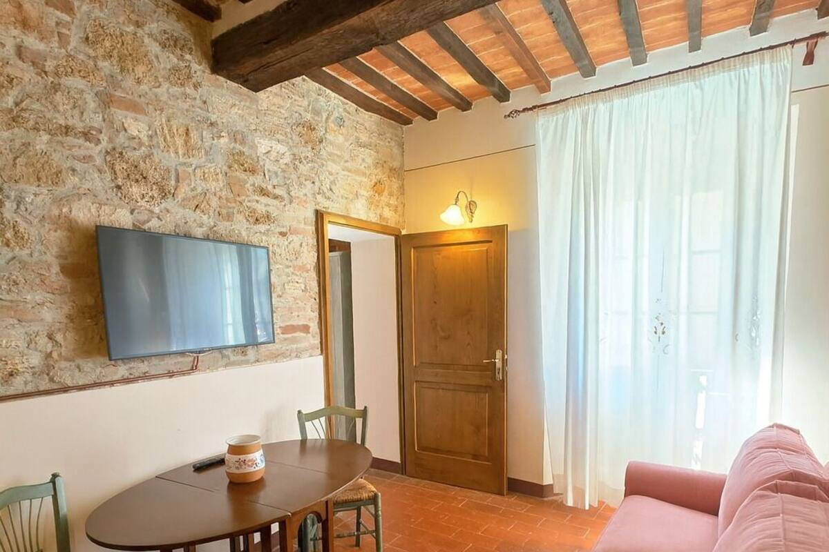 A cozy living room with a stone wall, wooden beams, a television, a dining table with chairs, and a pink sofa.