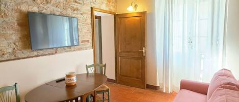 Una sala de estar acogedora con pared de piedra, vigas de madera, televisor, mesa de comedor con sillas y sofá rosa.