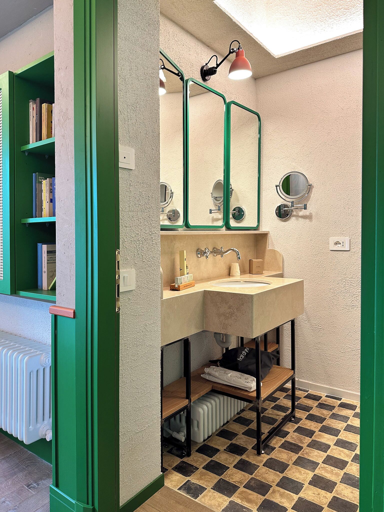 A bathroom with a checkered floor, a sink with a mirror, a radiator, and a green bookshelf.